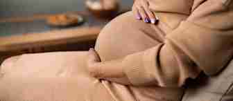 A pregnant person, wearing a beige dress, is sitting on a couch with one hand resting on their belly and the other hand placed below it. The background shows a wooden table with baskets and a plate. The person's nails are painted lavender.