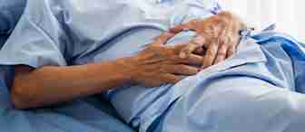 A person lying on a hospital bed wearing a light blue medical gown, with hands resting over their abdomen. The scene suggests a hospital or clinical setting with natural light filtering in from the background.