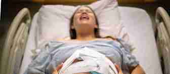 A pregnant woman lying in a hospital bed, gripping her belly with both hands. She has a medical monitor strapped to her abdomen and appears to be in labor, with an expression of pain or effort on her face. She is wearing a hospital gown.