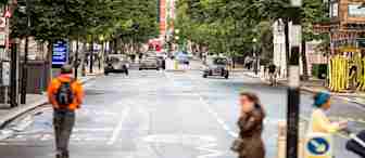 A city street scene with several cars and a taxi driving along. Pedestrians walk on sidewalks and cross the street, including a person in a bright orange jacket. Trees line both sides of the road, and buildings are visible in the background.
