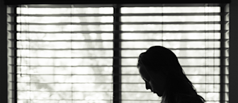 Black and white photo of a person with long hair standing in profile, facing right, and looking down. They are positioned in front of window blinds, with light filtering through, creating a contemplative or somber mood.