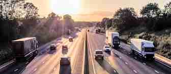 A busy highway at sunset with multiple lanes of traffic. Large trucks and cars are heading in both directions, surrounded by greenery and trees on either side. The sky is lit with a golden hue from the setting sun, casting long shadows on the road.