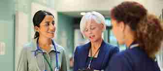 Three healthcare professionals stand in a hospital corridor, engaged in conversation. One has a stethoscope around her neck, another holds a clipboard, and the third is dressed in a nurse's uniform, holding a tablet.