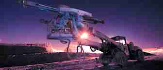 An industrial drilling machine with multiple attachments is illuminated against a dark, twilight sky. The ground appears excavated, and the machinery's silhouette is highlighted by a glowing light in the background.