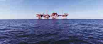 An offshore oil platform stands in the middle of the ocean against a backdrop of a clear, blue sky. The structure is supported by multiple stilts and has various cranes and equipment on top. The calm sea below reflects the purple hues of the platform.