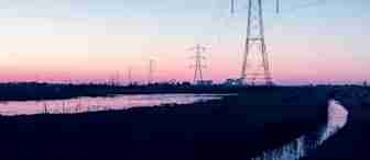 A series of electrical transmission towers and power lines stretch across a reflective water body at sunset. The sky transitions from a soft pink near the horizon to a serene blue higher up, with silhouettes of buildings and trees in the distant background.