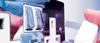 A close-up image of a person’s hand plugging a white UK three-pin plug into a shiny, metallic electrical socket on a white wall. The socket features dual switches, and their hand and the plug are reflected on the surface.