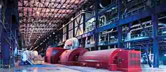 An industrial facility with large, red machinery and extensive metal piping. The space is open with a tiled floor and a high ceiling supported by metal beams. The background features a mix of pipes, ducts, and walkways, typical of a high-tech manufacturing plant.