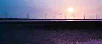 A serene landscape features numerous wind turbines on the horizon, silhouetted against a vivid sunset sky with hues of purple, pink, and blue. The turbines are reflected in a body of water below, with a dark and textured foreground adding depth to the scene.