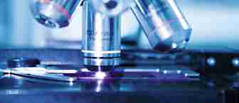 Close-up view of a microscope's objective lenses with detailed engravings visible, showing magnification levels. The image captures part of the slide stage and focus mechanisms. The background is softly blurred, focusing attention on the microscope.
