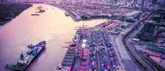 Aerial view of a large port with numerous shipping containers stacked neatly alongside large cranes. A river flows next to the port with a ship docked nearby. In the background, a sprawling city skyline is visible under a purple and pink sky.