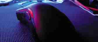 Close-up of a computer mouse on a dark mouse pad, illuminated by a purple and blue light. The background is slightly blurred but features a clutter of wires and computer components, with various LEDs and screens creating a moody, tech-centric atmosphere.