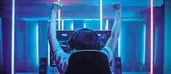 A person sits in a gaming chair with arms raised in victory, wearing headphones and facing a computer monitor with glowing neon lights in the background. The scene is illuminated with blue and purple hues, giving a futuristic and immersive gaming atmosphere.