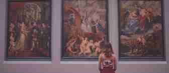 A person with shoulder-length hair, wearing a sleeveless top and skirt, sits on a bench in an art museum. They are facing three large classical paintings with intricate, vivid scenes, depicting numerous figures and actions in rich colors.
