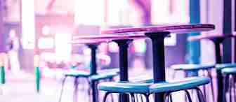 A close-up of tall bar stools and high tables in a modern indoor setting. The image is bathed in a purple and pink hue, creating a vibrant and stylish ambiance. The background is slightly blurred, emphasizing the seating area in the foreground.
