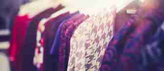 A row of colorful clothes hanging on white hangers in a brightly lit store. The garments feature various patterns, including houndstooth, floral, and solid colors. The focus is on the houndstooth-patterned garment in the foreground.