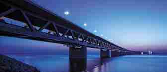 A long, illuminated bridge stretches across calm water at dusk. The sky transitions from deep blue to light blue at the horizon, with the bridge's lights reflecting on the water's surface. Rocks are visible in the foreground, forming a shoreline along the water.