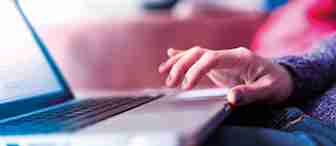 A close-up photo of a person using a laptop. The person’s right hand is typing on the keyboard. They have colorful nail polish and are wearing a long-sleeve sweater. The background is blurred, making it hard to discern specific details.