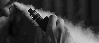 A close-up, black-and-white image of a person exhaling vapor from an e-cigarette, holding the device near their face. Dense vapor clouds are visible, and the person's mustache is slightly seen. The background is dark and blurry.