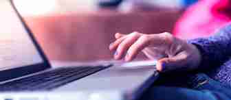 Close-up of a person's hand with purple nail polish resting on the trackpad of an open laptop. The background is softly blurred with pink and blue tones. The person appears to be using the laptop, which is situated on a flat surface.
