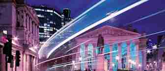 A nighttime city scene with light trails from passing vehicles, illuminating a historical building with columns and a statue in front. The sky is a deep blue, and the surrounding modern buildings are lit up, contrasting the neoclassical architecture.