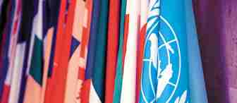 A collection of various national flags is displayed, prominently featuring the blue flag of the United Nations with its white emblem. The flags are closely arranged, showcasing a colorful and diverse array.