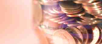 A glass jar filled with copper coins, mostly pennies, displayed against a softly blurred background. The coins reflect light, showing their shiny surfaces and engraved details. The jar is nearly full, with coins tightly packed inside.