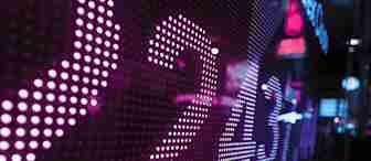 A close-up of a digital display with brightly lit numbers in various shades of purple and blue, possibly representing stock market data or financial information. The background is slightly out of focus, with blurry lights and neon-colored signs in an urban setting.
