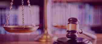 A wooden gavel rests on a sound block in the foreground, with a balanced scale of justice in the background. The setting appears to be a courtroom or a law library, indicated by the blurred shelves filled with books.
