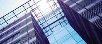 Looking up at a modern glass and steel office building with reflective windows. The sun is shining above, creating a bright, contrasting sky. The architectural design features clean lines, grid-like patterns, and a mix of metallic and glass surfaces.