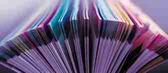 Close-up of the colorful, curved pages of an open book, fanned out to create a visually striking pattern. The pages feature various shades and hues, creating a vibrant and abstract appearance. The background is blurred in soft purple tones.