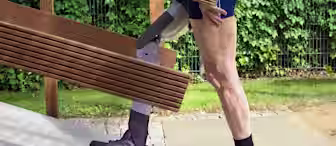 A person with a prosthetic leg is carrying wooden planks up a ramp outdoors. They are wearing black socks, black sneakers, and shorts, with a lush green hedge in the background. Their prosthetic limb is visible as they ascend the sloped surface.