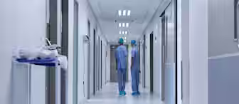 Two healthcare professionals in scrubs and caps stand in a hospital corridor, facing away. The hallway is brightly lit and lined with doors and medical equipment, creating a clean and professional atmosphere.