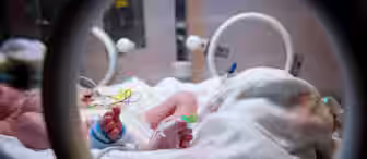 A newborn baby lies in an incubator, wrapped in a patterned blanket. The baby's feet are visible, with one foot adorned with a medical tag and an IV attached. Nearby medical equipment is partially visible, framing the scene.