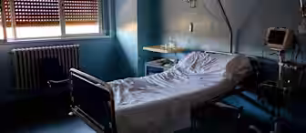 A dimly lit hospital room with a single unoccupied bed. The bed has white sheets and medical equipment nearby, including a monitor and an intravenous pole. Sunlight filters through the partially closed blinds. A chair and a radiator are positioned next to the window.