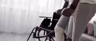 A person with a bandaged leg is seen using crutches to walk in a bright room. A wheelchair is positioned nearby, while a couch and sheer white curtains are visible in the background. The person appears to be wearing a white t-shirt and gray shorts.