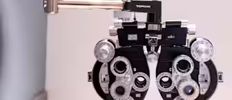 Close-up of an ophthalmic device, a phoropter, used for eye exams. It features numerous lenses, dials, and controls for assessing vision clarity. The word "Topcon" is visible on top of the device.