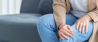 A person sits on a sofa, holding one knee with both hands. They are wearing blue jeans and a beige cardigan over a white shirt. The focus is on the hands and knee, suggesting discomfort or pain.