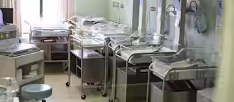 A hospital nursery room with several empty baby bassinets lined up. Each bassinet has a transparent cover and is equipped with basic monitoring devices. The room is softly lit, and curtains cover the windows partially.