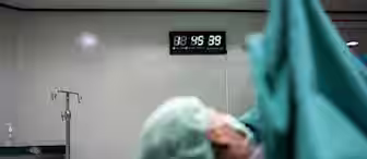 A patient lies on an operating table covered in green surgical drapes, with their face partially visible and a mask over their mouth. In the background, a wall-mounted digital clock displays the time as 8:56:39. Medical equipment and cabinets are also visible.