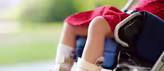 A child wearing a red dress sits in a wheelchair, with both feet in white orthopedic braces and resting on the footrests. The background is blurred and outdoors.