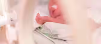 A newborn baby lies in an incubator, partially covered with a blanket. The baby's hand is visible, and a small tube is attached to their nose, indicating medical care. The focus is soft, with a warm, gentle tone.
