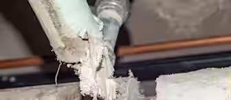 A close-up of a damaged and crumbling asbestos pipe in an industrial setting. The pipe appears to be deteriorating, with exposed sections showing fibrous asbestos material. The surrounding area has other pipes and metallic fixtures visible in the background.