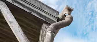Close-up of an old, weathered rain gutter and downspout attached to the corner of a wooden building, with a partly cloudy blue sky in the background.