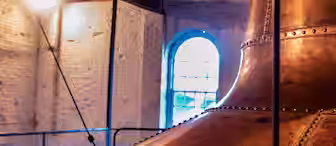 A large copper brewing kettle is seen inside a dimly lit brewery. The room has white brick walls and a large arched window in the background, illuminating the space with natural light. The ceiling is wooden with exposed beams.