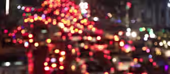 A blurred image of heavy traffic at night, showcasing numerous out-of-focus red and white lights from car tail lights and headlights, creating a bokeh effect. The overall scene gives a sense of congestion and movement.
