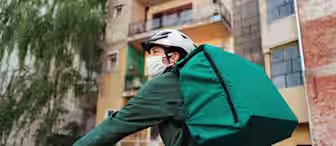 A food delivery cyclist wearing a mask and helmet rides in an urban area, carrying a large green backpack. Buildings with balconies are visible in the background, and the cyclist wears a green jacket.