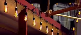A row of warm, hanging Edison-style light bulbs illuminated under a red wooden pergola. Green plants are in the foreground, and the background contains a blurred structure, all suggesting an outdoor setting, possibly a cozy patio or garden.