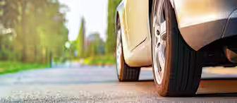 Close-up view of a car's tire on a sunlit road with blurred green trees and bushes in the background. The scene has a warm, serene atmosphere, and the road stretches out ahead.