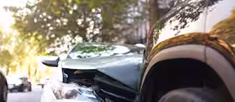 A silver car with a damaged front bumper has collided with the rear side of a black SUV on a city street lined with trees.